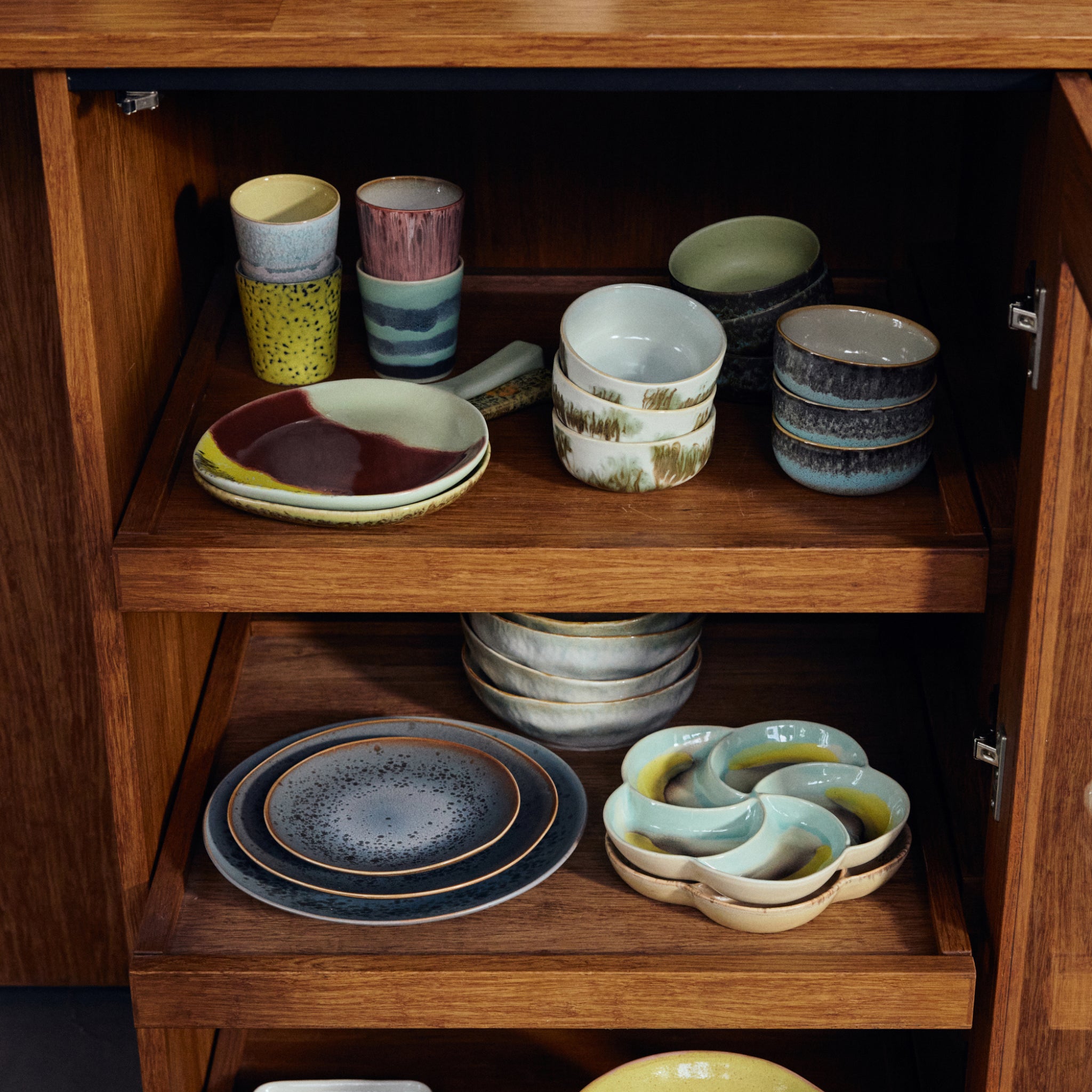 Een houten kast met drie planken toont gestapelde keramische kommen, mokken, kopjes en borden, waaronder een keramische serveerstuk en de HK Living 70s Ceramics Schotel Fjord in aardetinten, blauw gespikkelde en groene patronen.