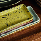 Een close-up van drie rechthoekige schotels gestapeld op hout, met de bovenste - een gespikkeld geel HK Living 70s Ceramics Serveerschaaltje XS Corn - rustend boven twee schotels in zachte pasteltinten.