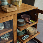 Een houten kast met glazen deuren toont gestapelde keramische borden en kommen, waaronder het HK Living 70s Ceramics Serveerschaaltje River, op twee uitschuifbare planken. Extra keramische serveerschaaltjes en houten voorwerpen rusten bovenop de kast.