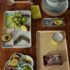 Een houten tafelset met HK Living 70s Ceramics Serveerschaaltje Patina, borden, kopjes en dienbladen bevat groene druiven, avocadohelften, kiwi, gemengde noten en een glas melk. Het HK Living servies heeft aardetinten en pastelkleuren.
