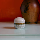 De HK Living 70s Ceramics Suikerpot Petal, een kleine ronde witte suikerpot met een gestructureerd oppervlak, staat op een lichtgroene ondergrond voor een grote bruine vaas en een oranje wand.