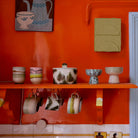 Een fel oranje keukenmuur heeft een plank met HK Living 70s Ceramics Tapas Kom op Voet Patina S, kopjes en kommen. Onder de plank hangen mokken, met rechts daarvan aardewerk, kunst en een blauw deurkozijn.