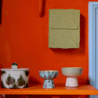 Drie HK Living 70s Ceramics Tapas Kom op Voet Patina S kommen, elk met hun eigen unieke vorm en kleur, staan op een plank tegen een feloranje muur, onder een lichtgroen kunstwerk en naast een lichtblauwe deurpost.