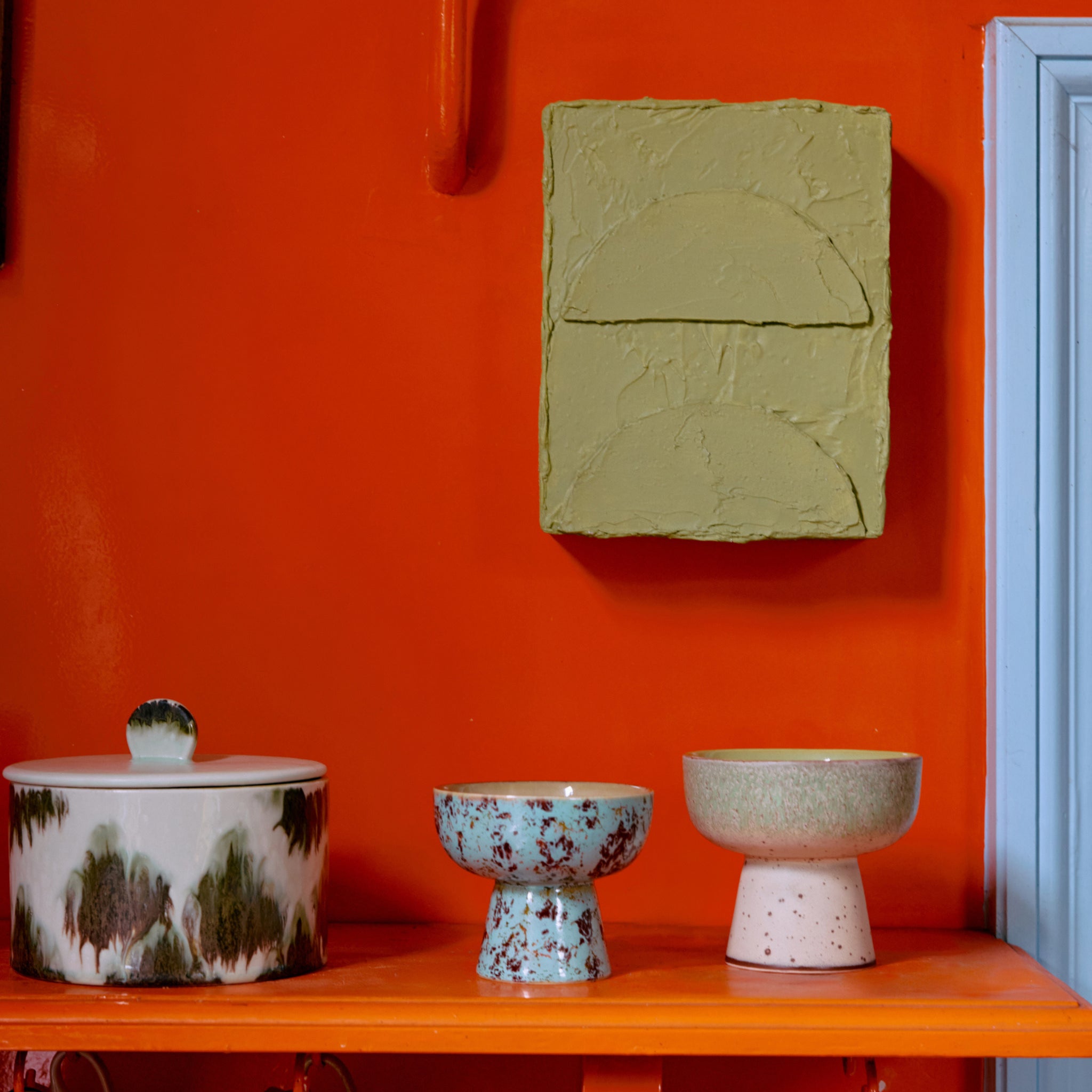 Drie HK Living 70s Ceramics Tapas Kom op Voet Patina S kommen, elk met hun eigen unieke vorm en kleur, staan op een plank tegen een feloranje muur, onder een lichtgroen kunstwerk en naast een lichtblauwe deurpost.