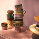 Een stapel kleurrijke HK Living 70s Ceramics kopjes en kommen staat op een roze oppervlak in het zonlicht. Vlakbij staan een mok, een hoge pot en de HK Living 70s Ceramics Tapas Kom op Voet Lush L met de gele rand gedeeltelijk zichtbaar aan de rechterkant.