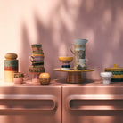 Een collectie kleurrijke HK Living 70s Keramiek, waaronder de Tapas Kom op Voet Lush L, is gerangschikt op een blush roze kast tegen een bijpassende muur, met zacht zonlicht en bladerrijke schaduwen die een warme, uitnodigende scène creëren.