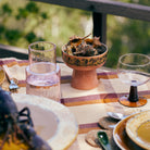 Een gedekte tafel buiten met de HK Living 70s Ceramics Tapas Kom op Voet L, glazen en een schaal met schelpen op een gestreept tafelkleed. Zonlicht benadrukt het glaswerk en keramiek, met groen op de achtergrond.