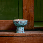 De HK Living 70s Ceramics Tapas Kom op Voet Patina S, met een blauw en bruin gevlekte afwerking, staat op een houten oppervlak bij een houten raam met een groene muur op de achtergrond.