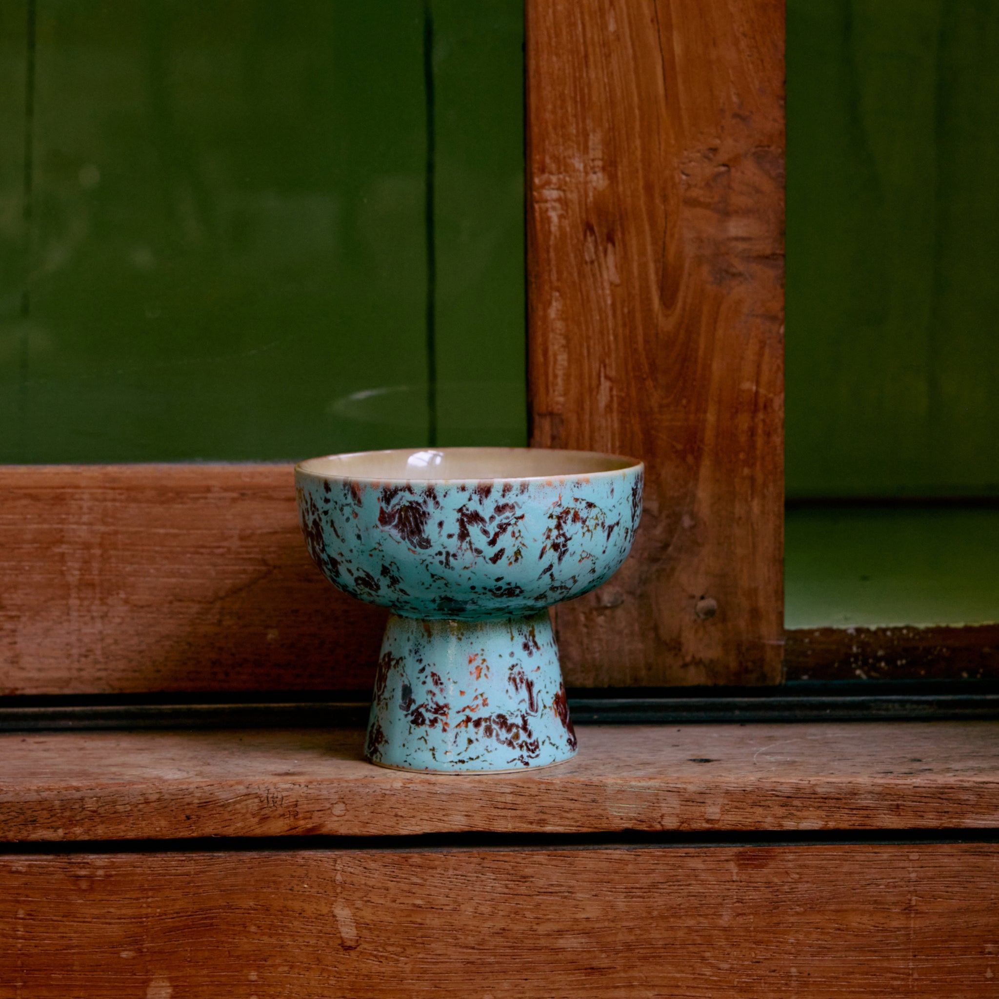 De HK Living 70s Ceramics Tapas Kom op Voet Patina S, met een blauw en bruin gevlekte afwerking, staat op een houten oppervlak bij een houten raam met een groene muur op de achtergrond.