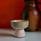 De HK Living 70s Ceramics Tapas Kom op Voet Slush L, met een gespikkelde gebroken witte basis en een kom met groene textuur, staat op een licht oppervlak. Daarachter staat een rustieke bruine kan tegen een oranje muur.