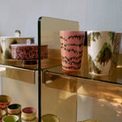 Op glazen planken met gouden steunen tegen een bleke muur tonen HK Living 70s Ceramics Theekop barokke kopjes en andere keramische schalen roze en groen gemarmerde patronen.