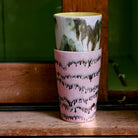 Twee HK Living 70s Ceramics Theekop Baroque bekers zijn gestapeld op een houten oppervlak. Het bovenste kopje heeft groene en bruine druppels; het onderste heeft roze met zwarte golvende motieven. Een houten paneel en groene achtergrond maken het tafereel compleet.