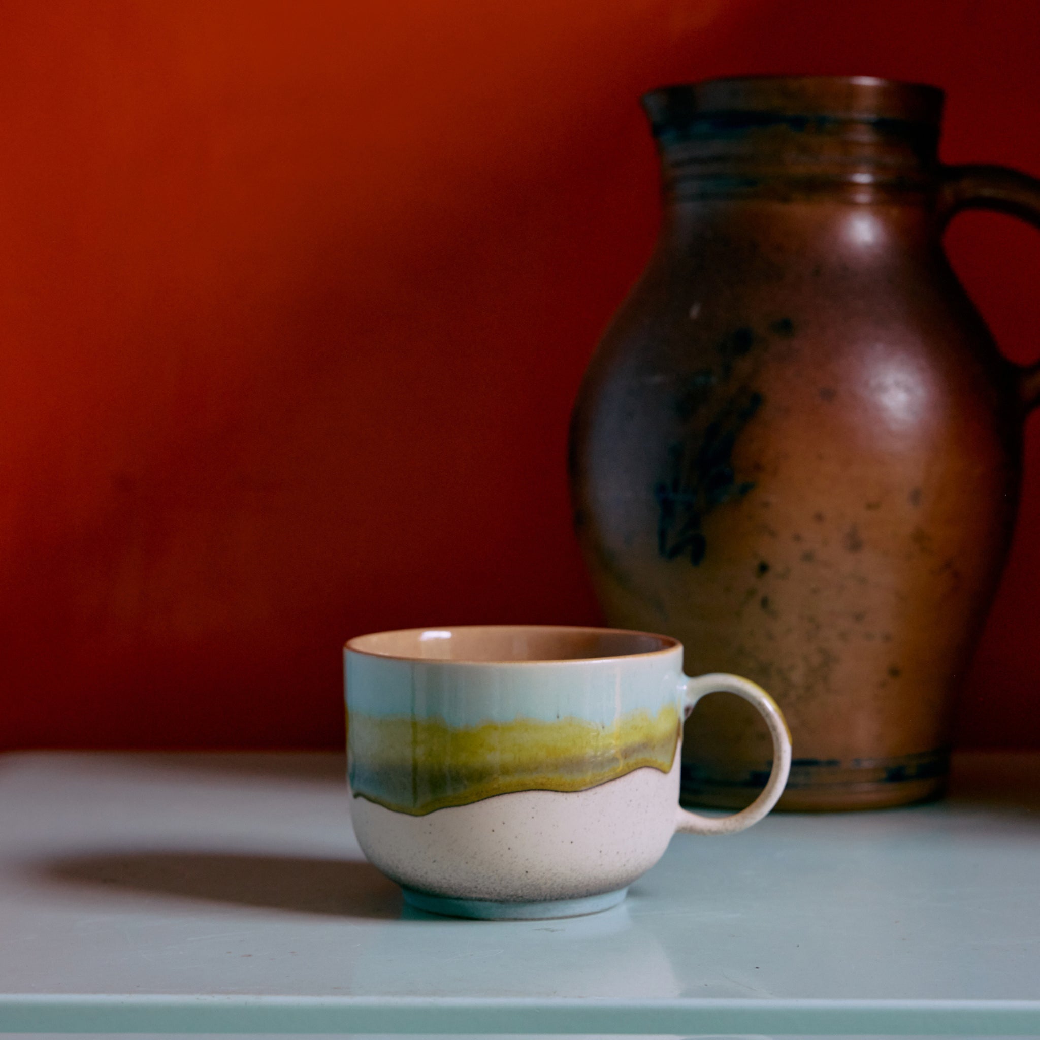 De HK Living 70s Ceramics Theekop Glint, met groen en beige glazuur, staat op een lichtblauw oppervlak voor een rustieke bruine kan en oranje achtergrond, die de vintage stijl van HK Living oproept.