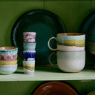 Op een plank staan HK Living 70s Ceramics XS Kom Modernist 4 st., gestapelde kopjes, modernistische kommen met patronen en twee grote mokken - allemaal gerangschikt voor een groene achtergrond met decoratieve borden.