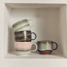 Vier HK Living 70s Ceramics Theekop Quest mokken staan gestapeld op een witte verzonken plank. Drie met pastelkleurig en aards glazuur staan op elkaar gestapeld, terwijl er één rechtop naast de groep staat.