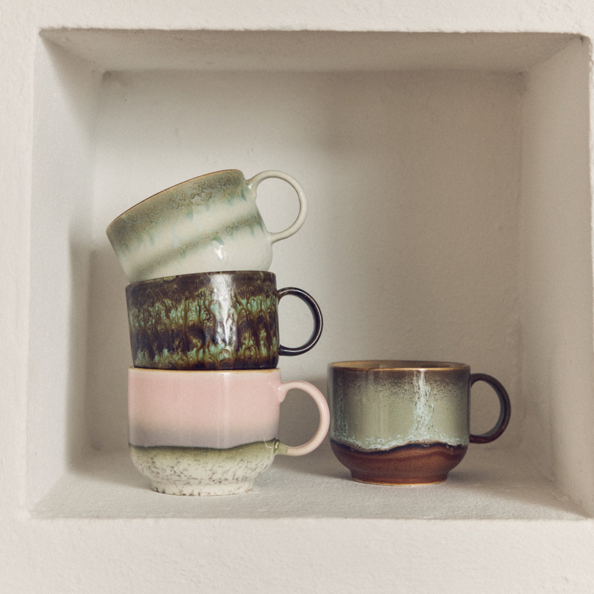 Vier HK Living 70s Ceramics Theekop Quest mokken staan gestapeld op een witte verzonken plank. Drie met pastelkleurig en aards glazuur staan op elkaar gestapeld, terwijl er één rechtop naast de groep staat.