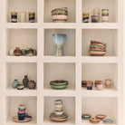 Kleurrijke keramische kommen, kopjes, borden en de HK Living 70s Ceramics Vaasjes Pursuit (5 st.) van HK Living zijn gerangschikt op een witte ingebouwde plank met drie rijen-ideaal voor retro interieur decoratie. Elke nis benadrukt unieke stukken aardewerk.