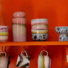Een levendige oranje plank toont kleurrijke HK Living keramische kopjes, kommen en een 70s Ceramics Voorraadpot Glazuur in pasteltinten. Vier mokken hangen aan haken onder de plank.