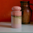 De HK Living 70s Ceramics Voorraadpot Glazuur in roze, met een ronde deksel, rust op een licht oppervlak met een bruine kan erachter en een oranje muur op de achtergrond.