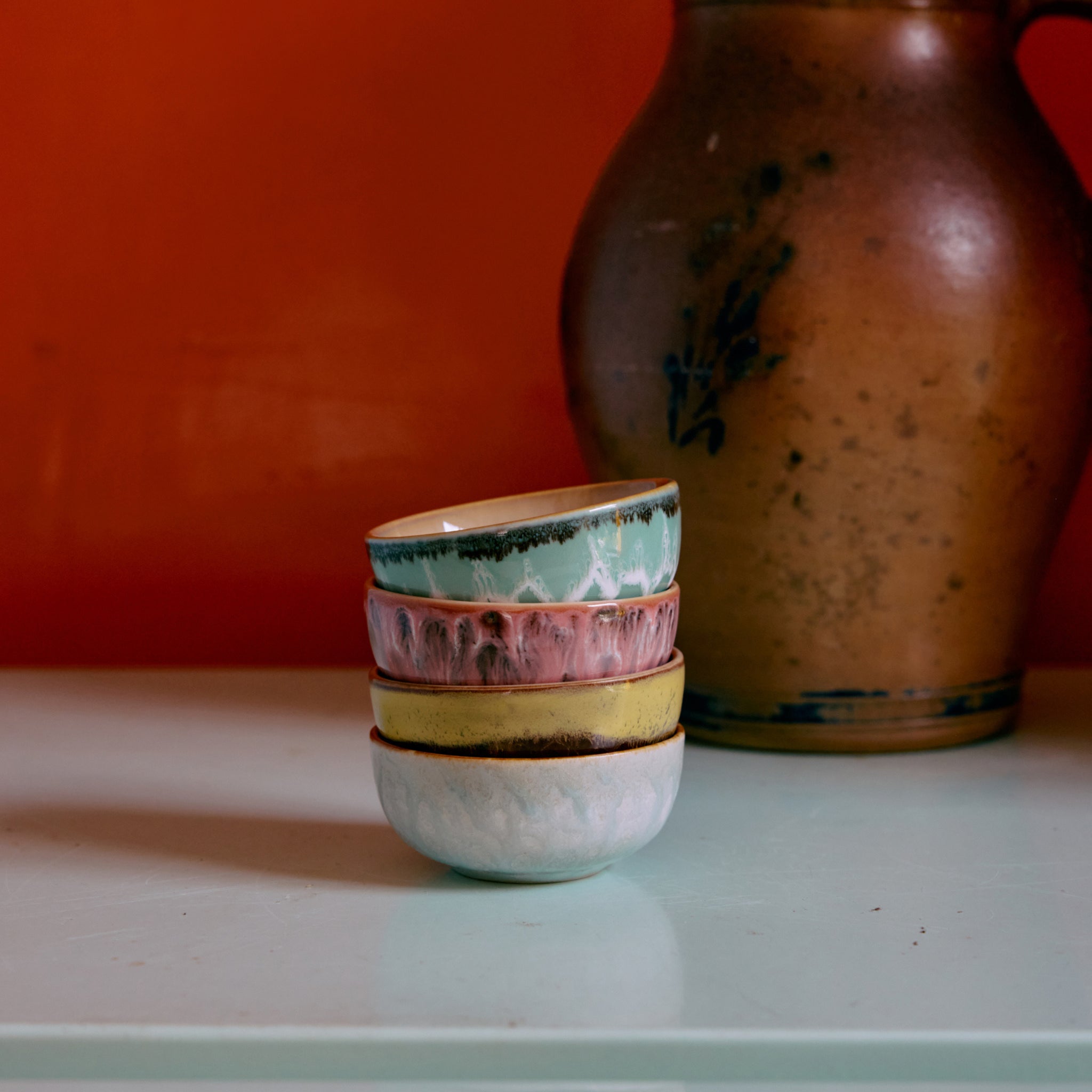 Vier HK Living 70s Ceramics XS Kom Modernist kommen zijn gestapeld op een lichtblauw oppervlak, met een grote bruine vaas op de achtergrond tegen een oranje muur - ideaal voor fans van de kenmerkende stijl van HK Living.