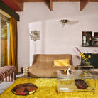 Een retro woonkamer met een bruine bank, geel kussen en glazen salontafel op een felgeel vloerkleed. De HK Living Allure Bijzettafel Crème voegt charme toe, gecombineerd met een gestreepte poef, moderne muurkunst en lange gele gordijnen.