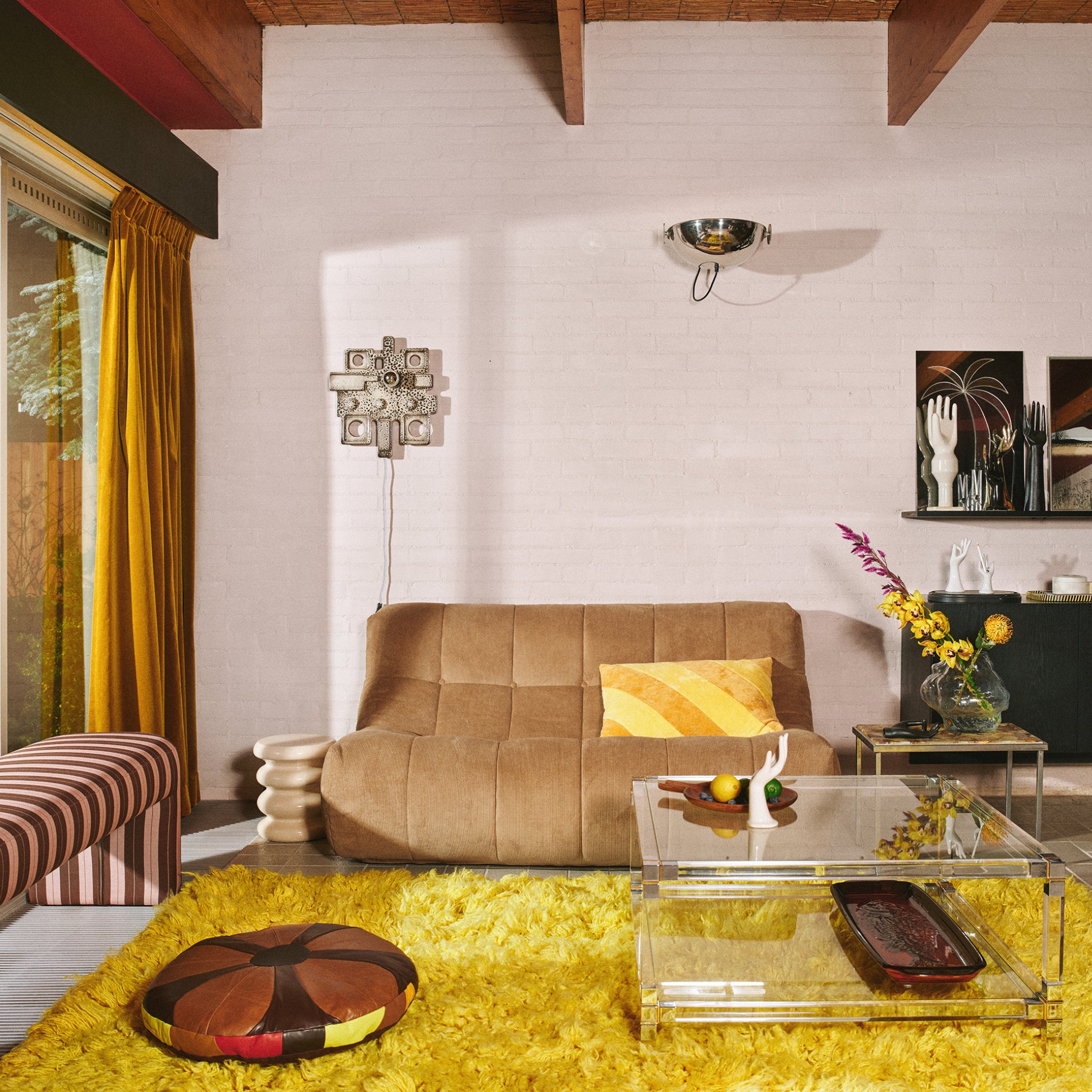 Een retro woonkamer met een bruine bank, geel kussen en glazen salontafel op een felgeel vloerkleed. De HK Living Allure Bijzettafel Crème voegt charme toe, gecombineerd met een gestreepte poef, moderne muurkunst en lange gele gordijnen.