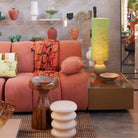 Een moderne woonkamer met een roze sofa, kleurrijke kussens, houten en witte krukjes, een groene lamp en de HK Living Allure Bijzettafel Crème te midden van decoratieve planken.