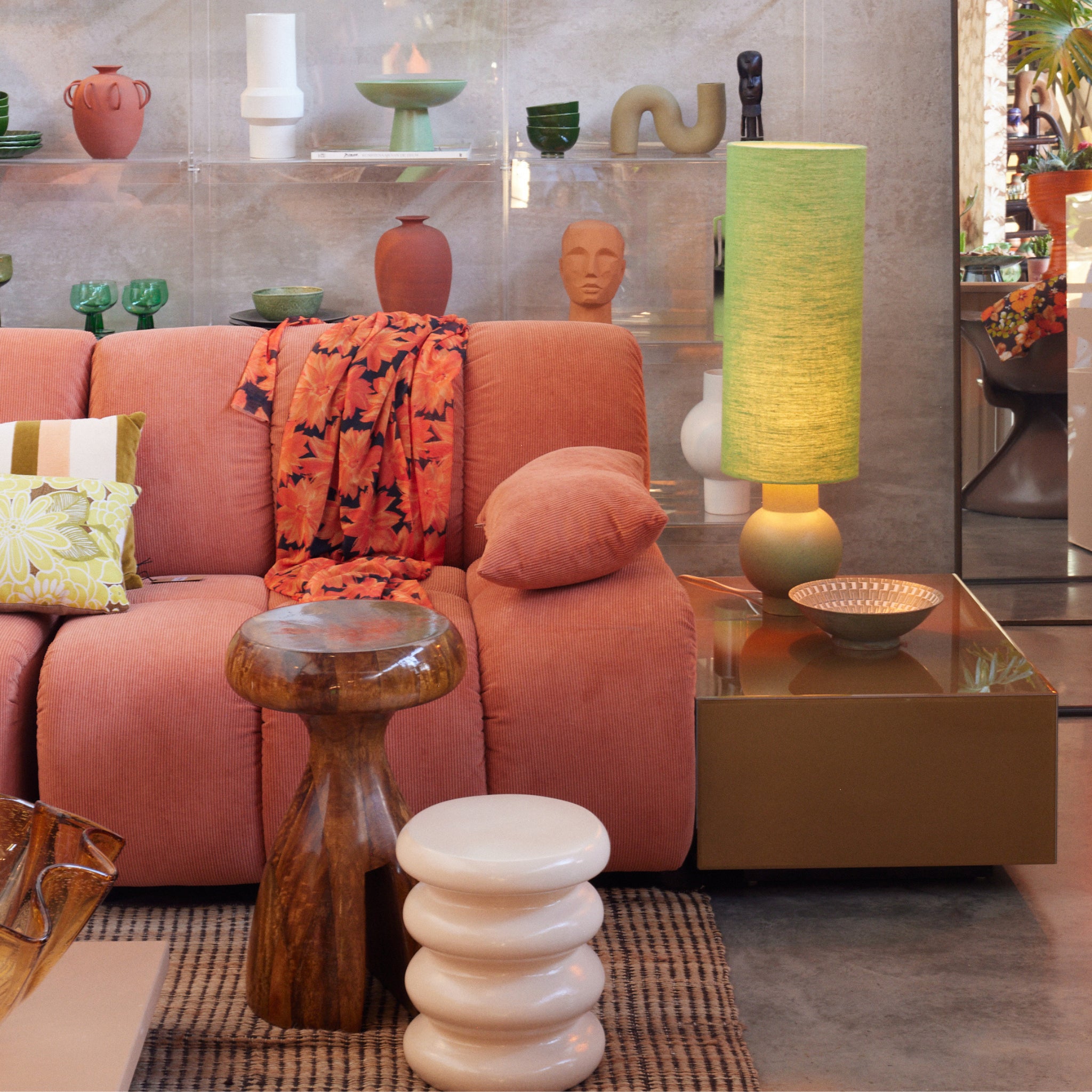 Een moderne woonkamer met een roze sofa, kleurrijke kussens, houten en witte krukjes, een groene lamp en de HK Living Allure Bijzettafel Crème te midden van decoratieve planken.