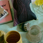 Een close-up van een sierkussen met een golvend gestikt patroon, omringd door HK Living Artist Ceramics Koffiekop 4 st., een klein gestreept kopje, een roze boek en een gele bloem op een lichtgroene ondergrond.