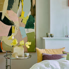 Een moderne slaapkamerhoek met een kleurrijk abstract schilderij, een kleine ronde tafel met een witte vaas met gele bloemen, een HK Living Artist Ceramics Koffiekop 4 st. en pastelkleurige kussens op het bed naast een witte schouw.