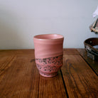 Een HK Living Artist Ceramics Koffiemok uit de 4-delige set, met een roze gestructureerde, gespikkelde onderkant, staat op een houten tafel tegen een lichte muur, met vage objecten op de achtergrond rechts.