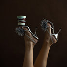 Twee voeten in zilveren sandalen met hoge hakken en metallic pompons steken af tegen een donkere achtergrond. Eén hak balanceert een HK Living Christmas Special Geurkaars Frosted Leaves kaars.