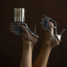 Twee voeten, met glanzend zilveren sandalen met hoge hakken en glinsterende pompons van klatergoud, worden in de lucht geheven. Op één schoen staat een HK Living Christmas Special Geurkaars Peperkoekkaars. De achtergrond is donkerbruin.