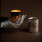 Een gehandschoende hand houdt een brandende kaars vast in een bruine keramieken houder met zwarte druppels. Ernaast staat een HK Living Christmas Special Geurkaars Gingerbread kaars in een metalen houder. De achtergrond is donkerbruin.