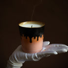 Een witgehandschoende hand houdt de HK Living Kerstspecial Geurkaars Peperkoekkaars vast, met een roze en zwart druppelontwerp. De lont is net gedoofd, met een rookpluimpje tegen een donkerbruine achtergrond.