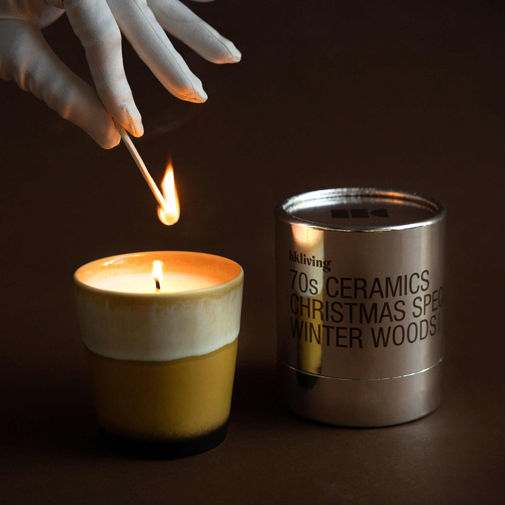 Een gehandschoende hand steekt een kaars aan terwijl de HK Living Kerstspecial Geurkaars Winterbos, in een metalen houder, ernaast staat. De donkerbruine achtergrond voegt warmte toe en benadrukt het gezellige HK Living tafereel.
