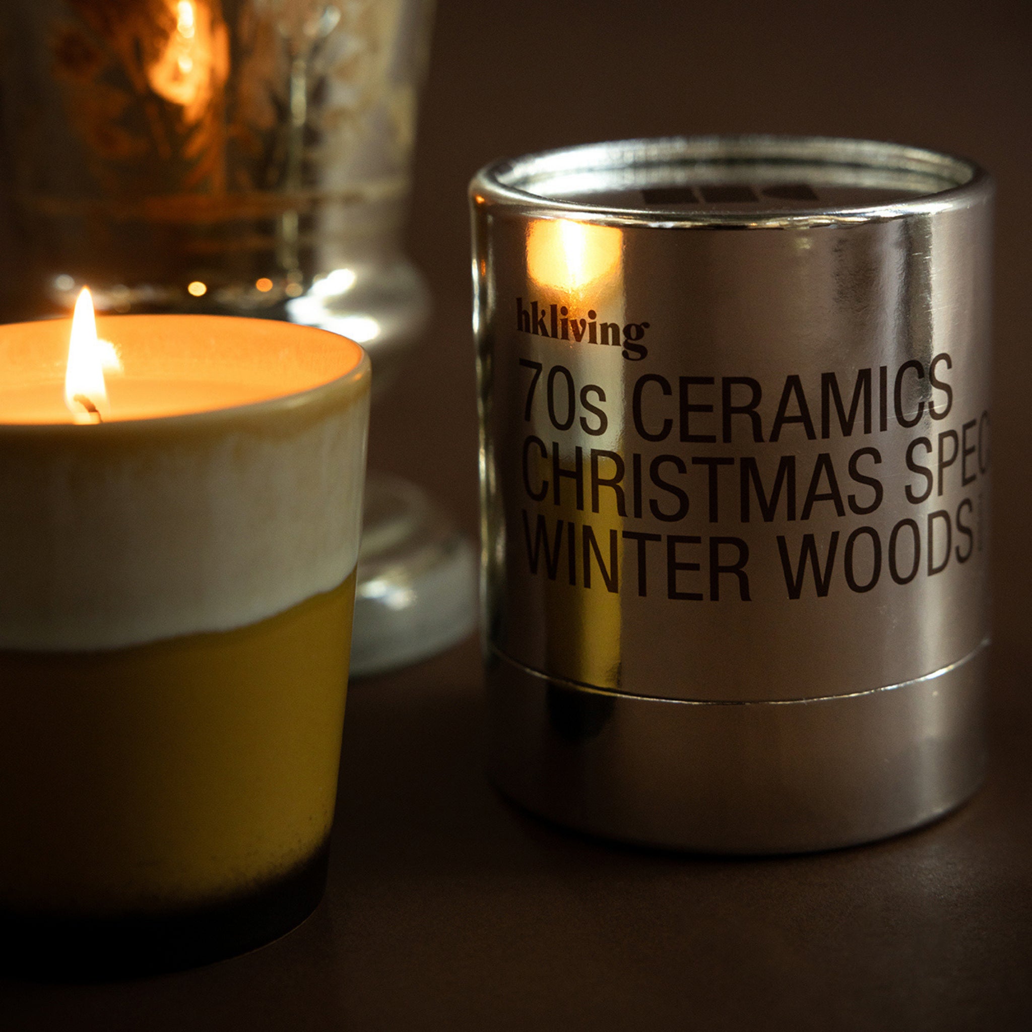 Een brandende kaars in een geel en crèmekleurige houder staat naast een HK Living Christmas Special Geurkaars Winter Woods metalen blik op een donker oppervlak, met een decoratief glas en 70s Ceramics koffiemok op de achtergrond.