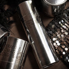 De HK Living Kerstspecial Geurkaarsen Frost & Firewood 3 st. in zilver metallic potjes met zwarte tekst worden getoond naast zwarte veren en een pailletten accessoire-een stijlvol kerstcadeau of om je favoriete geurkaarsen te presenteren.