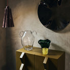 Een stilleven met een gestructureerde vaas van helder glas, een HK Living Coralla Theelichthouder Lime kom op een mosterdkleurige kast, een kastanjebruine hanglamp en een grote zwarte wandsculptuur tegen een grijze muur.