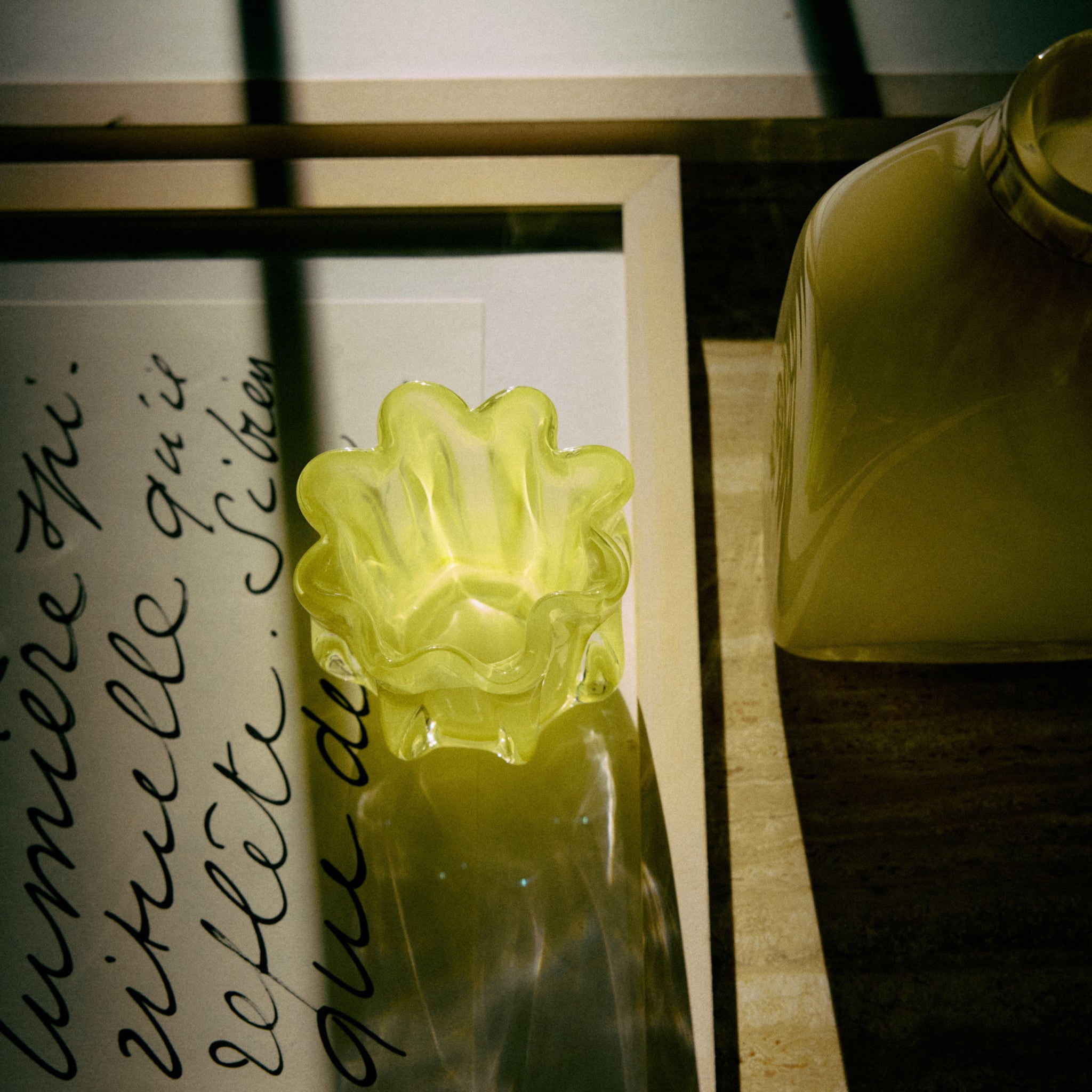 De HK Living Coralla Theelichthouder Lime, met zijn golvende rand en gele tint, werpt schaduwen op een ingelijst handgeschreven briefje en gele vaas op hout in warm, gedempt zonlicht.