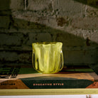 Een HK Living Coralla Theelichthouder Lime met een golvend ontwerp ligt op gestapelde boeken, waaronder "EVOCATIVE STYLE", tegen een rustieke bakstenen muur in zachte, stemmige verlichting.