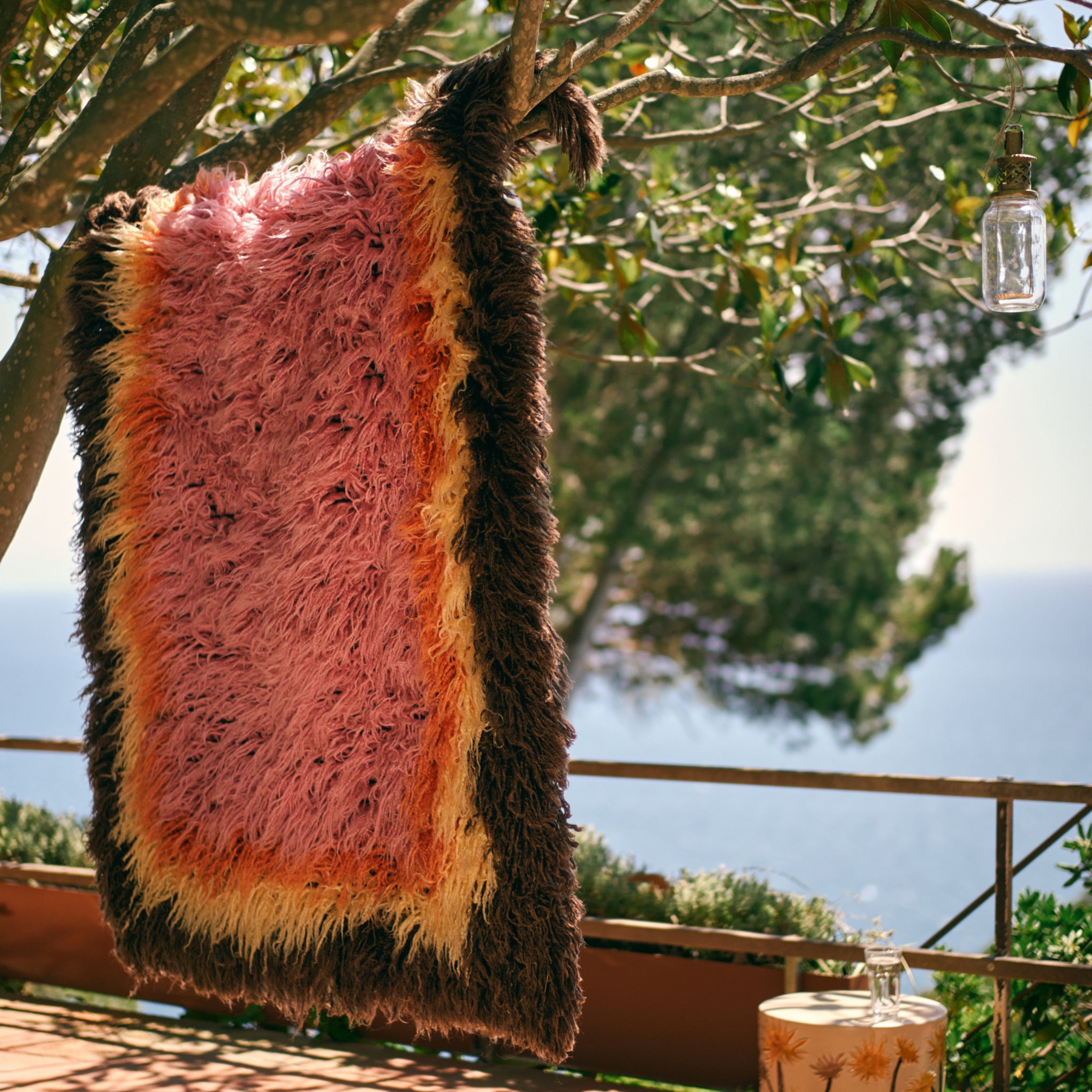 De HK Living Fluffy Tapijt Downtown 180/120, met levendige roze, gele, oranje en bruine strepen, hangt buiten aan een boomtak aan zee en geeft charme aan een tafel met een glazen pot. Perfect als stijlvol vloerkleed.