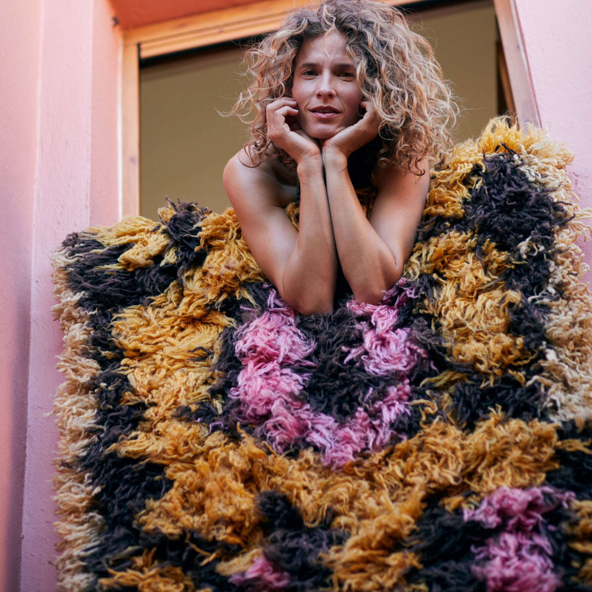 Een vrouw met krullend haar glimlacht terwijl ze op een vensterbank leunt, haar armen rustend op de HK Living Fluffy Tapijt South Beach 360/80, met gele, roze en zwarte patronen. Roze muren en een open raam maken het vrolijke tafereel compleet.