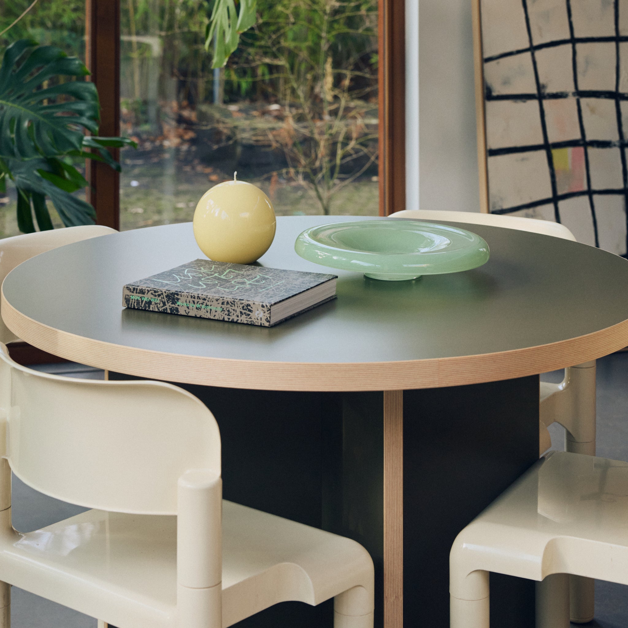 Op een ronde tafel met een groen blad liggen een notitieblok, een gele bolvormige kaars en de HK Living Gallery Serveerschaal Mint D34 als designaccent, omringd door witte stoelen. Grote planten en een raam staan op de achtergrond.