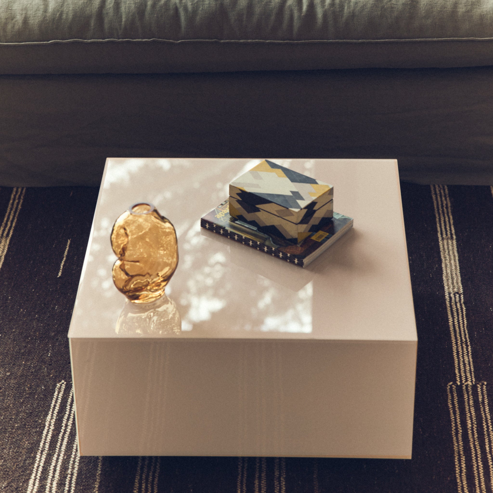 Op een wit glanzende salontafel staan een HK Living Gleam Vaas Amber H18, een doos met geometrisch patroon en een boek. De tafel staat op een donker gestreept tapijt met een grijze bank erachter, terwijl het zonlicht gevlekte schaduwen werpt over de ruimte.