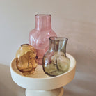 De HK Living Gleam Vaas Roze H32 staat samen met amberkleurige en grijze abstracte golvende vazen op een ronde crèmekleurige sokkel tegen een neutrale achtergrond.