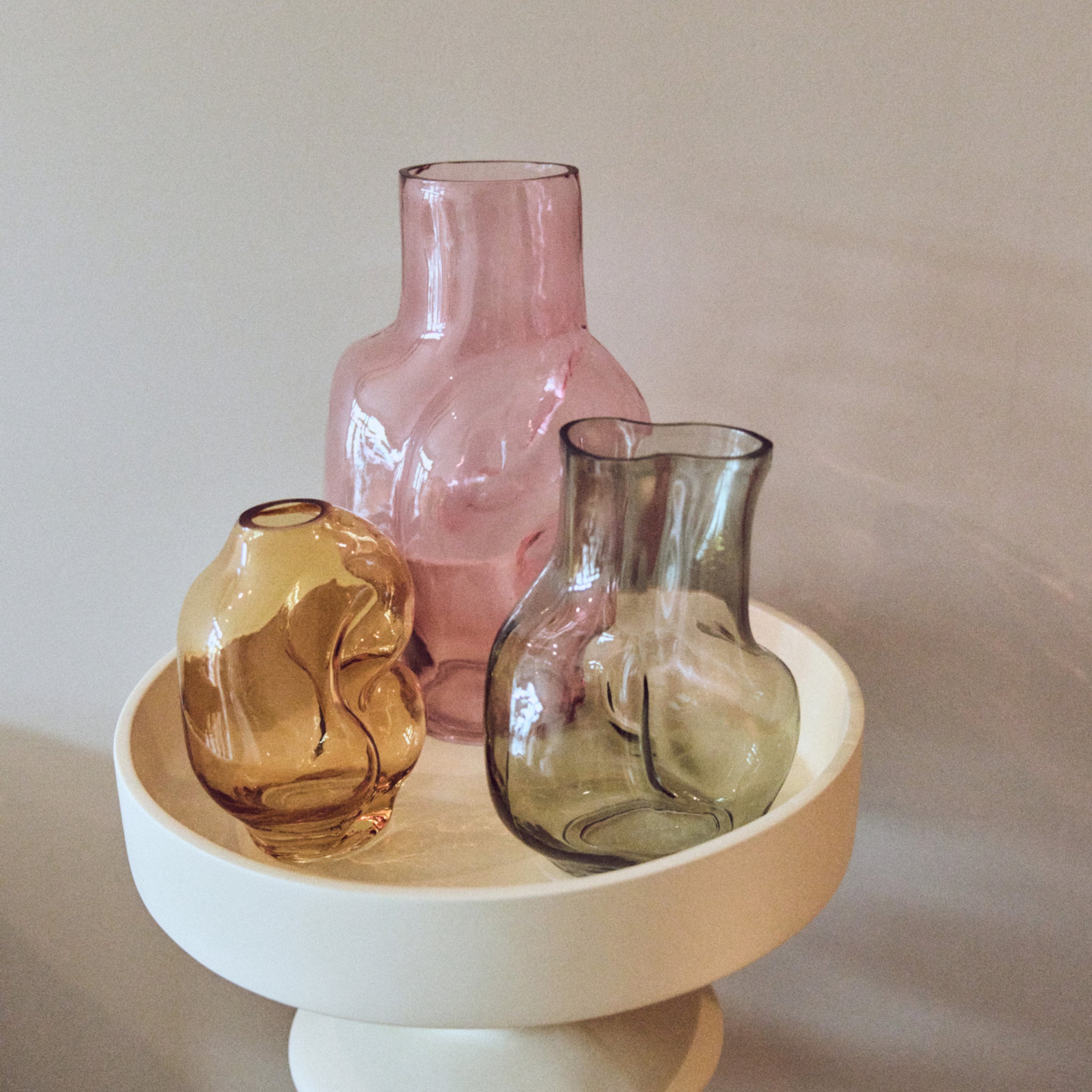 De HK Living Gleam Vaas Roze H32 staat samen met amberkleurige en grijze abstracte golvende vazen op een ronde crèmekleurige sokkel tegen een neutrale achtergrond.