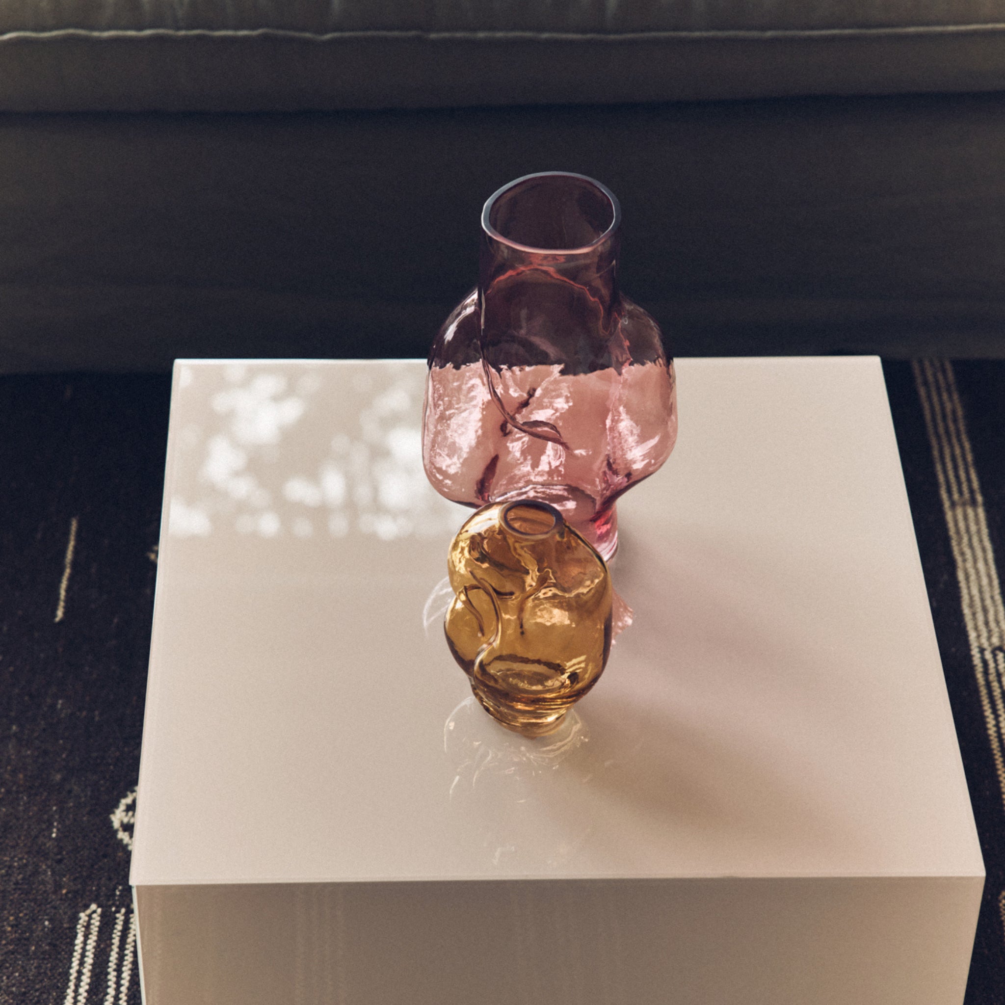 Een HK Living Gleam Vaas Roze H32 en een kleine amberkleurige glazen vaas rusten op een glanzend witte vierkante tafel in zonlicht, met een donkere bank en een vloerkleed met patroon gedeeltelijk zichtbaar op de achtergrond.