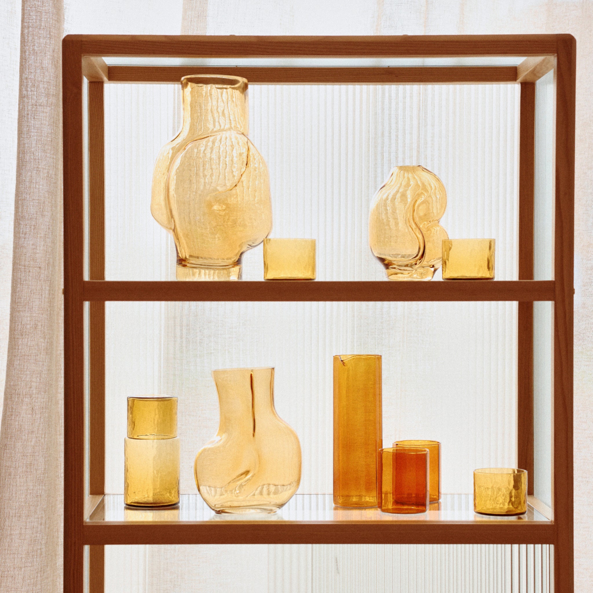 Op een houten plank staat de HK Living Gleam Vaas Amber H18 van HK Living, samen met andere glazen vazen in verschillende vormen en maten, tegen een lichte, doorschijnende achtergrond.