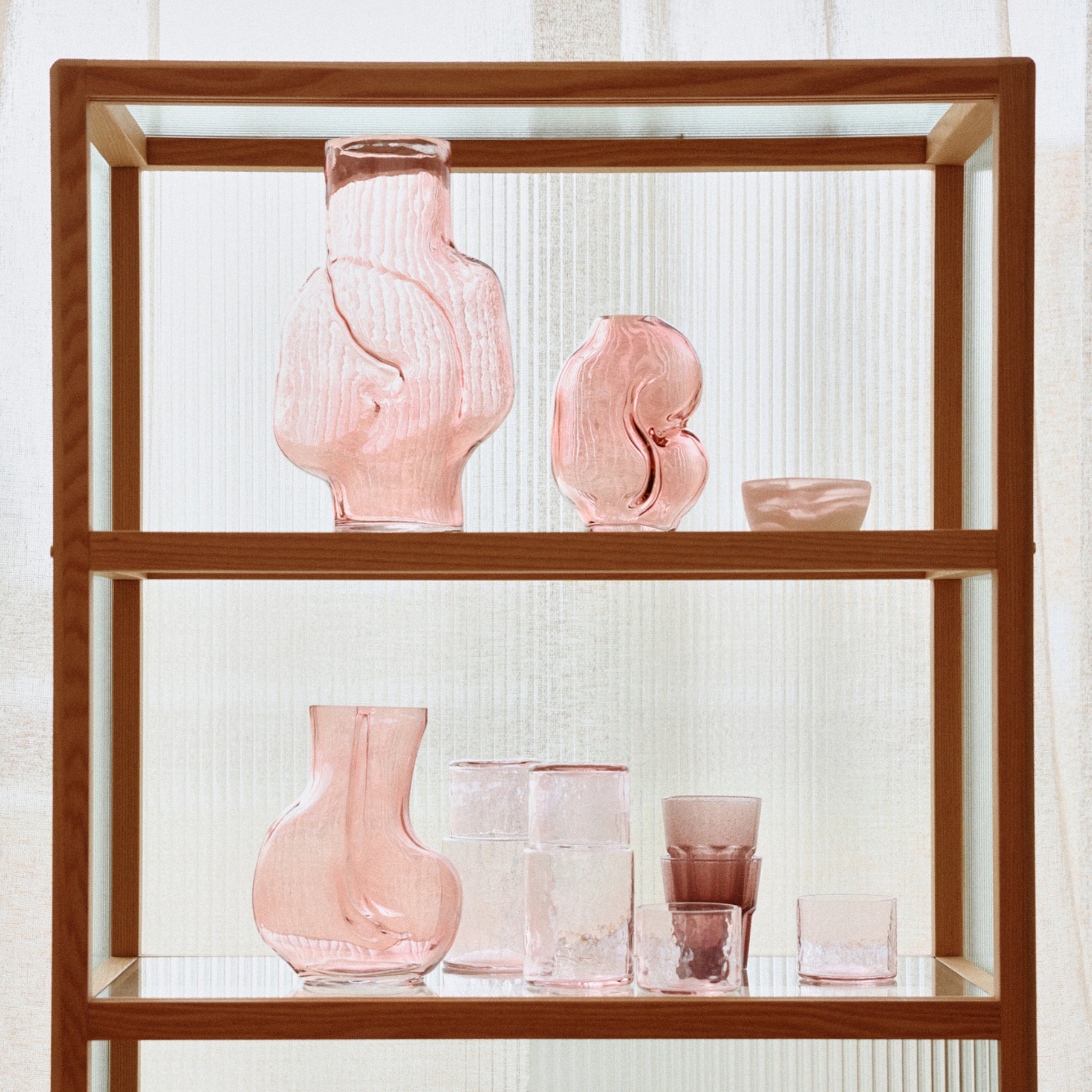 Op een houten plank staan de HK Living Gleam Vaas Roze H32 en glazen kopjes met een heldere structuur in verschillende abstracte vormen tegen matglazen panelen.